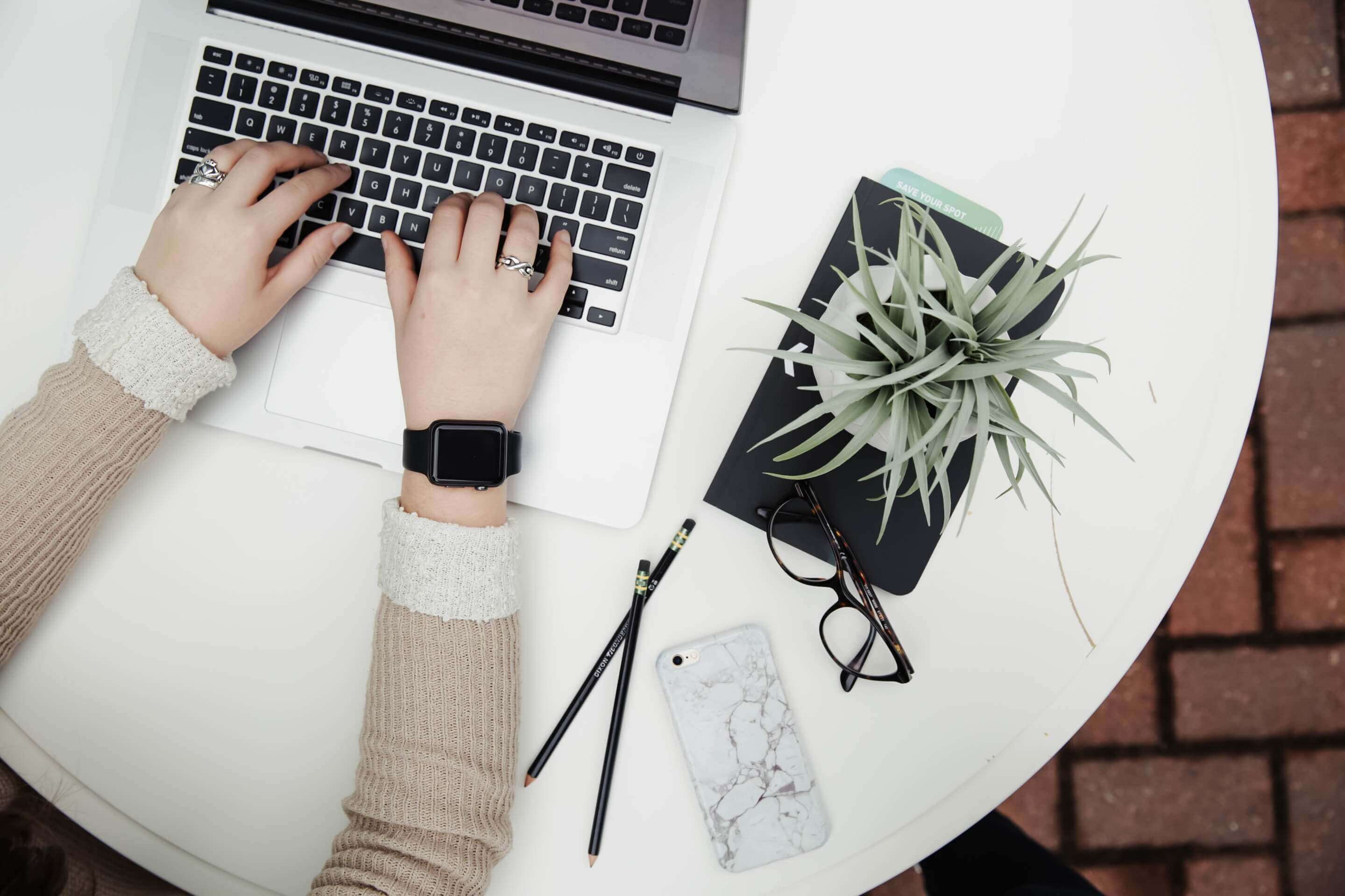 Hands of a person on a laptop, which stands on a white, round table. Next to the laptop is a plant standing on a book.