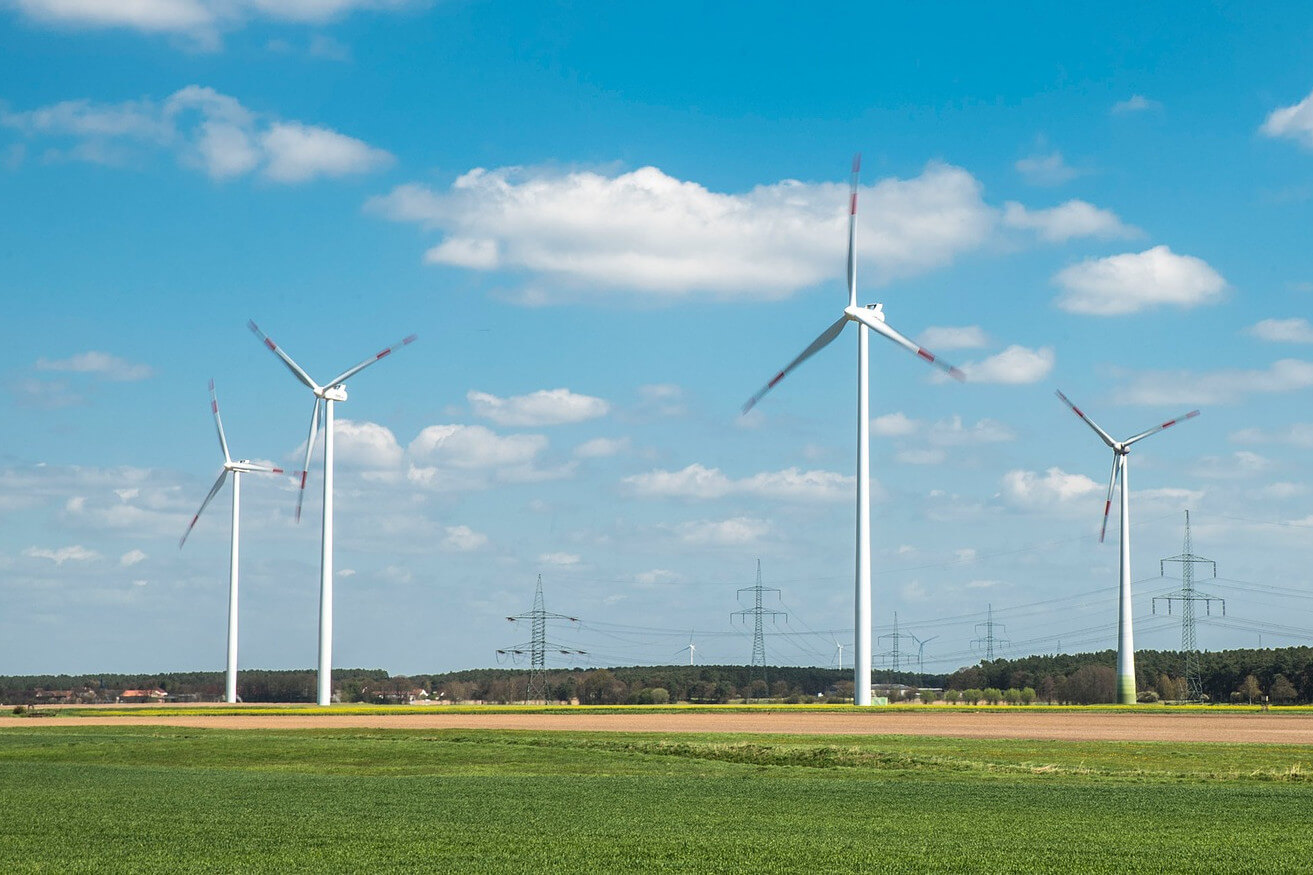 Windkrafträder auf einer grünen Wiese. Im Hintergrund eine Stromtrasse und Berge