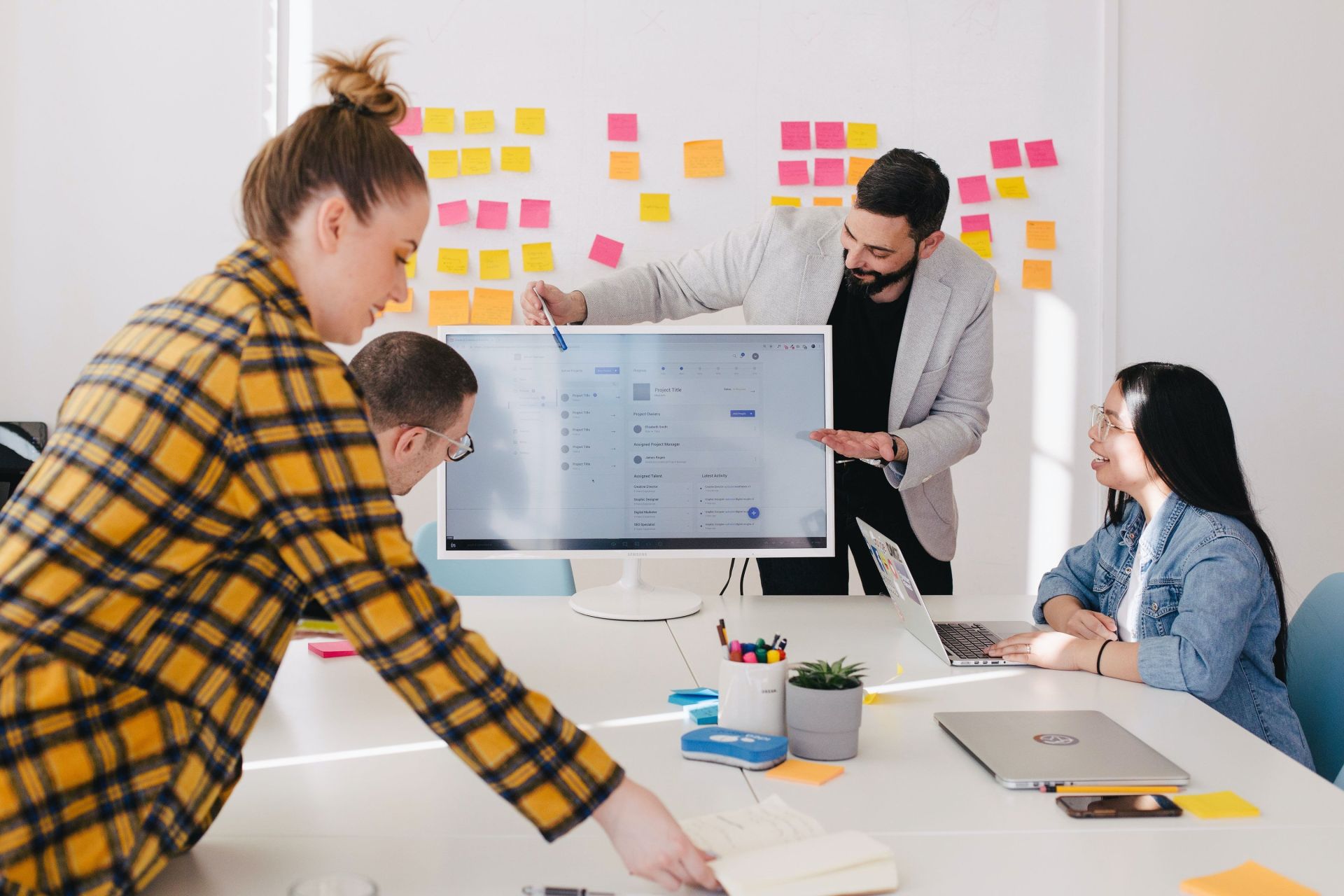 4 people at a table, in the background post-its on the wall with notes. On the table is a monitor with a website on which one of the people explains certain contents with a pen.